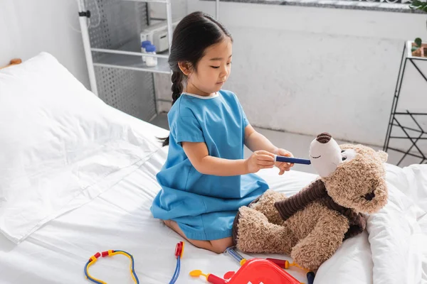 Азиатка в больничном халате держит игрушечный шприц рядом с плюшевым мишкой во время игры на кровати в клинике — стоковое фото