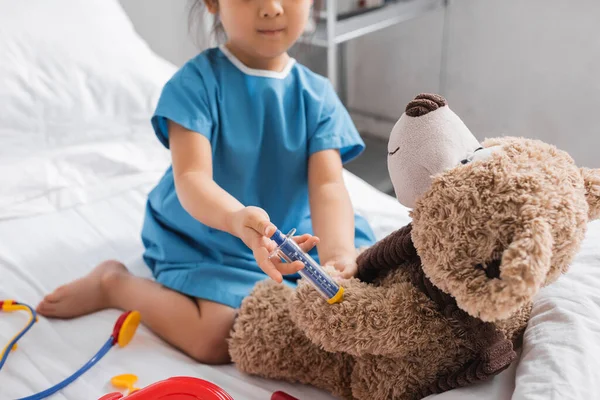 Partial view of child with toy syringe doing injection to teddy bear on hospital bed — Stock Photo