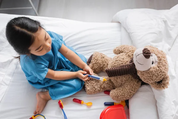 Top view of asian child playing on hospital bed and doing injection to teddy bear with toy syringe — Stock Photo