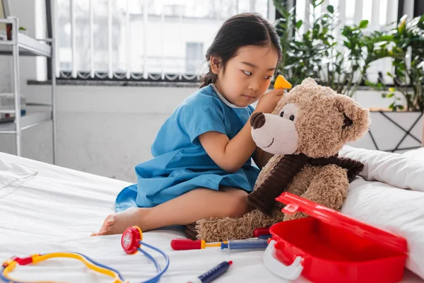 Asian child in hospital gown examining teddy bear with toy otoscope on bed in pediatric clinic — Stock Photo