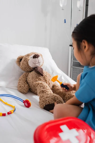 Borrosa asiático niño examinando teddy oso con reflex martillo en hospital cama - foto de stock