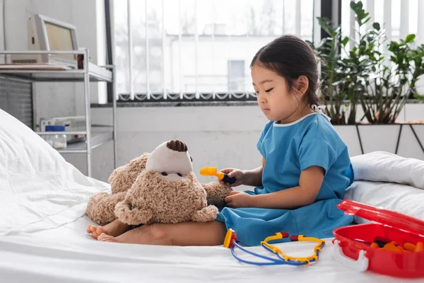 Vista lateral de poco asiático chica examinando teddy oso con juguete reflex martillo en hospital sala - foto de stock