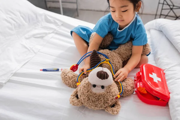 Asiatisches Mädchen legt Spielzeug-Stethoskop auf Teddybär, während sie auf Bett in Kinderklinik spielt — Stockfoto
