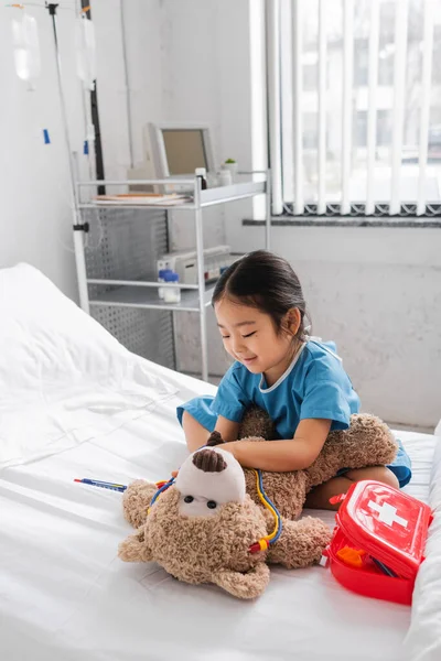 Cheerful asian girl playing with toy stethoscope and teddy bear on bed in hospital ward — Stock Photo