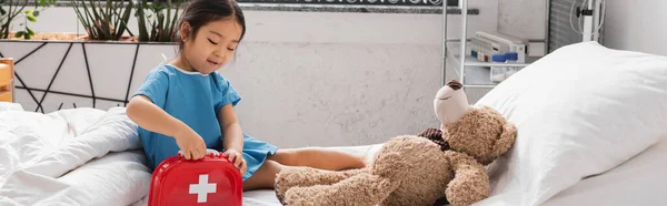 Asian child opening toy first aid kid near teddy bear on hospital bed, banner — Stock Photo