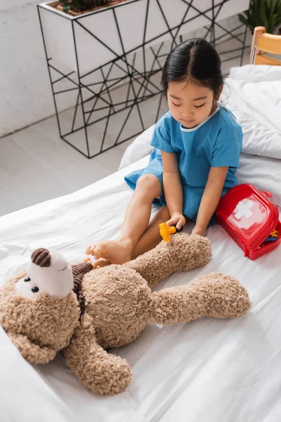 Vue grand angle d'un enfant asiatique jouant sur un lit d'hôpital et examinant un ours en peluche avec un marteau réflexe jouet — Photo de stock