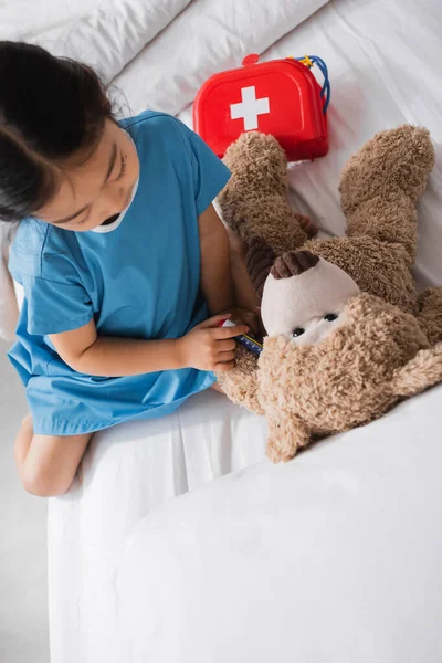 Вид сверху на азиатского ребенка с игрушечным шприцем, делающего инъекцию плюшевому мишке во время игры в больничном отделении — стоковое фото