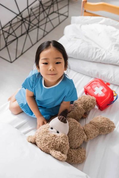 Vista superior da menina asiática sorrindo para a câmera enquanto brincava com ursinho de pelúcia e equipamentos médicos de brinquedo na cama do hospital — Fotografia de Stock