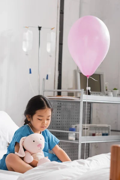Pouco asiático criança brincando com brinquedo coelho enquanto sentado no hospital cama perto festivo balão — Fotografia de Stock