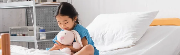 Smiling asian girl embracing toy bunny while sitting on bed in hospital ward, banner — Stock Photo