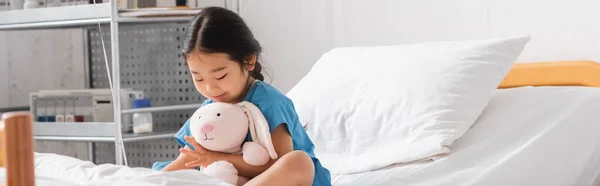 Lächelnde asiatische Mädchen umarmen Spielzeughasen, während sie auf dem Bett in der Krankenstation sitzen, Banner — Stockfoto