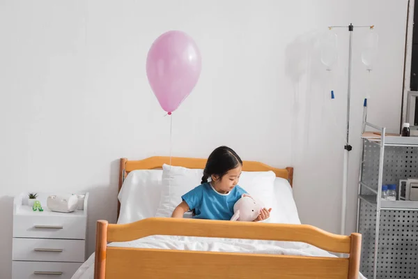 Little asian girl playing with toy bunny on bed under festive balloon in hospital ward — Stock Photo