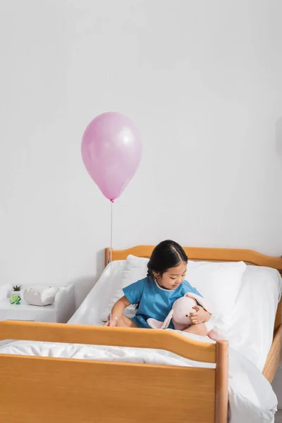 Asian kid in hospital gown sitting on bed with soft toy under festive balloon — Stock Photo