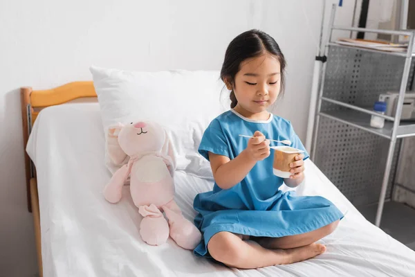 Pleased asian girl holding tasty yogurt while sitting on hospital bed near toy bunny — Stock Photo