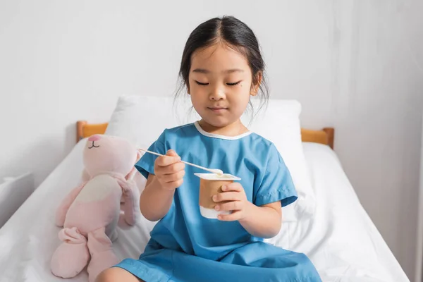 Piccola ragazza asiatica in abito da ospedale mangiare delizioso yogurt durante la colazione in clinica — Foto stock
