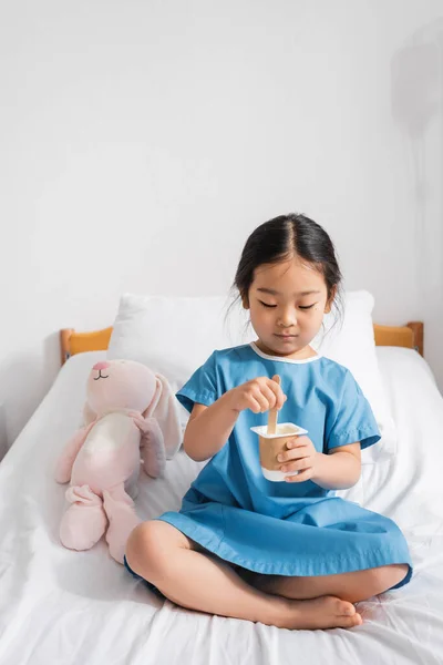 Босоногая азиатская девушка смешивает вкусный йогурт, сидя рядом с игрушечным кроликом на больничной койке — стоковое фото
