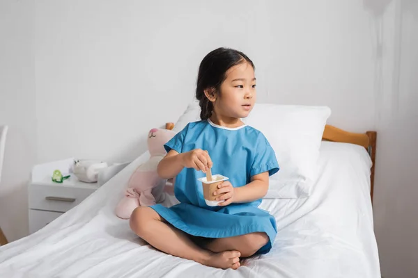 Menina asiática em vestido de hospital olhando para longe enquanto comendo iogurte saboroso na cama na clínica pediátrica — Fotografia de Stock