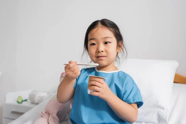 Positive asian child in hospital gown eating delicious yogurt in clinic — Stock Photo