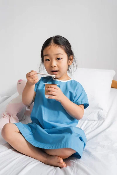 Menina asiática comer delicioso iogurte e olhando para a câmera durante o café da manhã no hospital — Fotografia de Stock