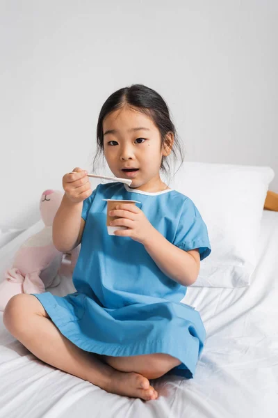 Asian girl eating delicious yogurt and looking at camera during breakfast in hospital — Stock Photo
