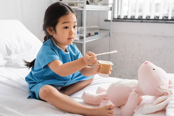 Barefoot asian girl holding delicious yogurt near toy bunny while sitting on hospital bed — Stock Photo