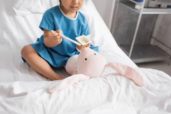 Cropped view of girl in hospital gown feeding toy bunny with yogurt on bed in clinic — Stock Photo