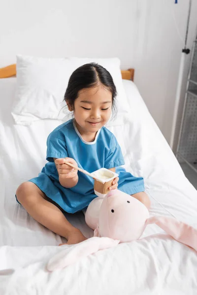Cheerful asian girl feeding soft toy with yogurt while playing in hospital ward — Stock Photo