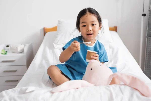 Feliz asiático criança olhando para câmera e comer saboroso iogurte perto de soft toy no hospital cama — Fotografia de Stock
