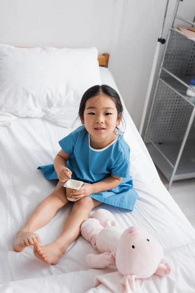 Positive asian girl holding delicious yogurt and smiling at camera in hospital ward — Stock Photo