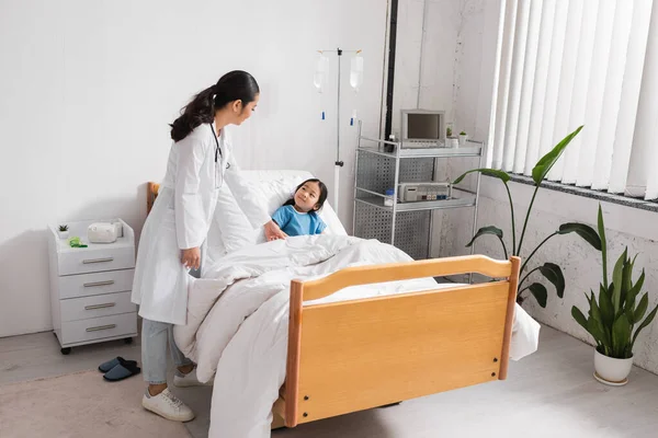 Pediatra no branco casaco olhando positivo asiático menina no moderno hospital ward — Fotografia de Stock