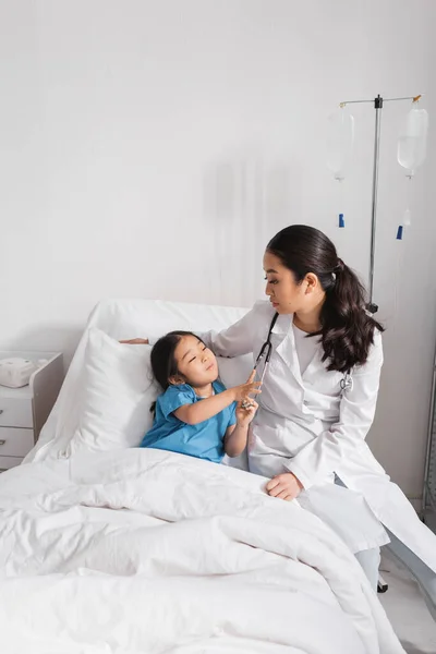 Любопытная азиатка, трогающая стетоскоп рядом с доктором в больнице — стоковое фото