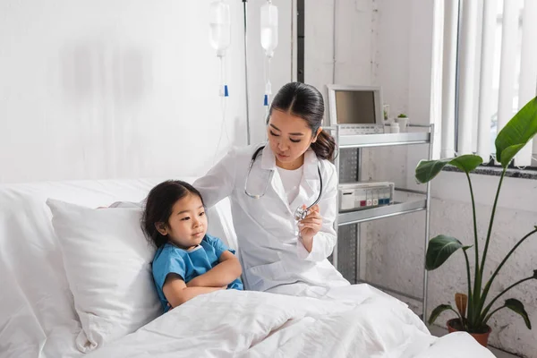Junger Arzt zeigt asiatischem Mädchen Stethoskop, das mit verschränkten Armen auf dem Krankenhausbett sitzt — Stockfoto