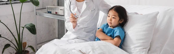 Asian girl crossing arms near doctor with stethoscope in pediatric clinic, banner — Stock Photo