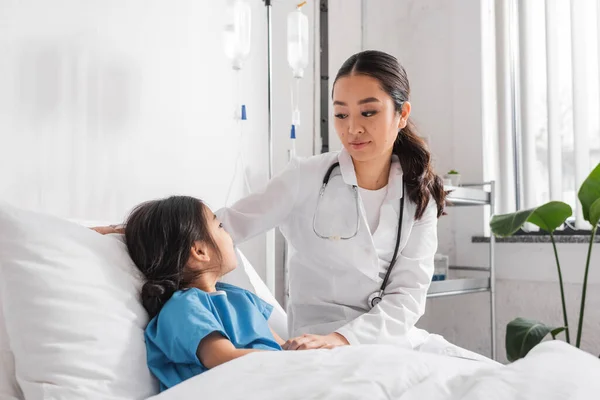 Young asian pediatrician with stethoscope looking at girl on bed in hospital ward — Stock Photo