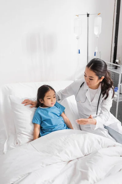 Asiático pediatra mostrando mano a poco chica sentado en hospital cama - foto de stock