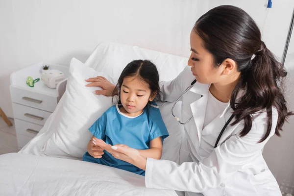 Pequeña chica asiática mirando a mano de médico mientras está acostado en cama de hospital - foto de stock