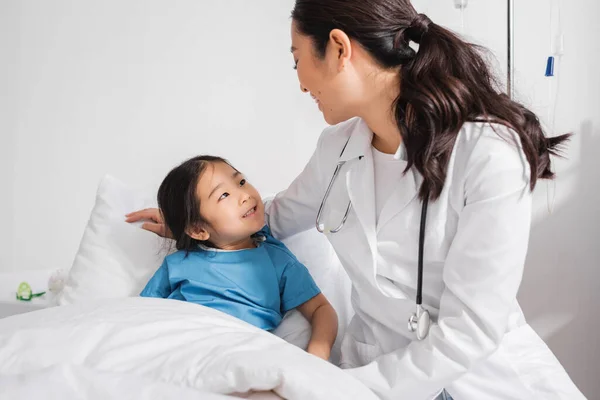 Беззаботная азиатская девушка и доктор в белом халате улыбаются друг другу в больничном отделении — стоковое фото