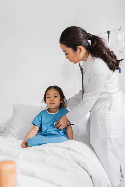 Молодой педиатр осматривает азиатского ребенка со стетоскопом на кровати в клинике — стоковое фото