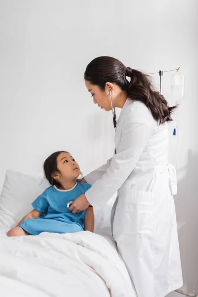Asian kid sitting on hospital bed and looking at pediatrician with stethoscope — Stock Photo