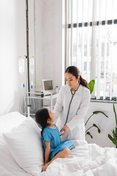 Asiático pediatra con estetoscopio examinar poco paciente sentado en hospital cama - foto de stock