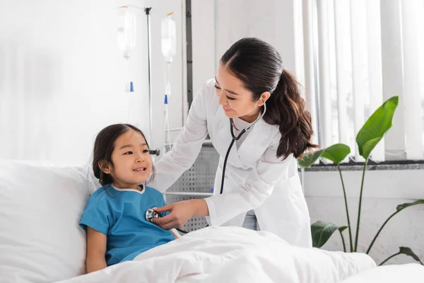 Joven asiático médico con estetoscopio examinar alegre chica en pediátrico hospital - foto de stock