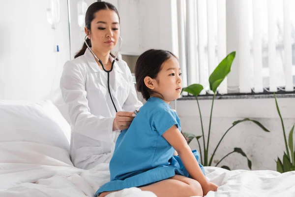 Atento médico examinando asiático chica en hospital bata sentado en cama en clínica - foto de stock