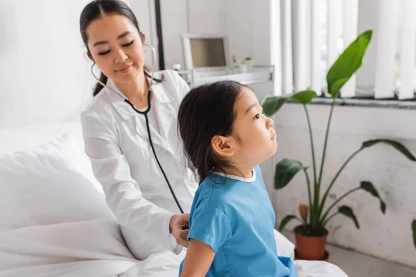Smiling asian doctor with stethoscope examining little patient sitting on hospital bed — Stock Photo