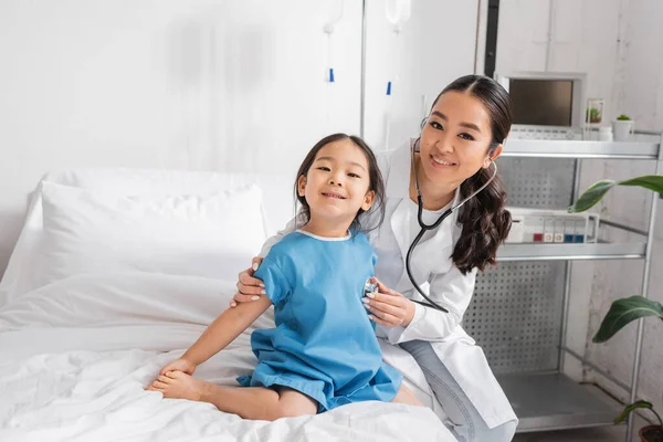 Sonriente asiático doctor con estetoscopio mirando cámara mientras examinar pequeño niña en pediátrico clínica - foto de stock