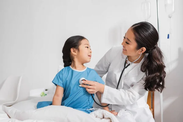 Médico positivo em casaco branco examinando menina asiática com estetoscópio em hospital ward — Fotografia de Stock