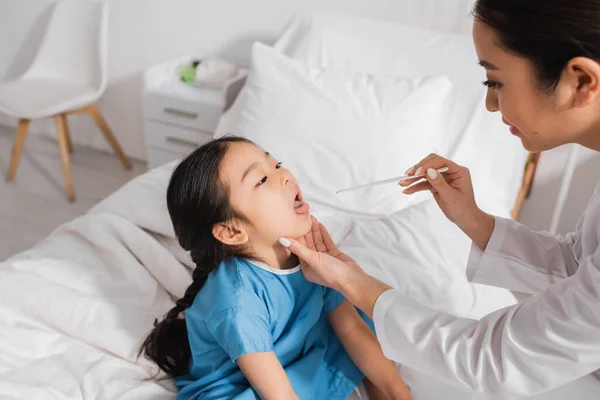 Asiático chica sentado con abierto boca cerca pediatra con lengua depresor en hospital sala - foto de stock