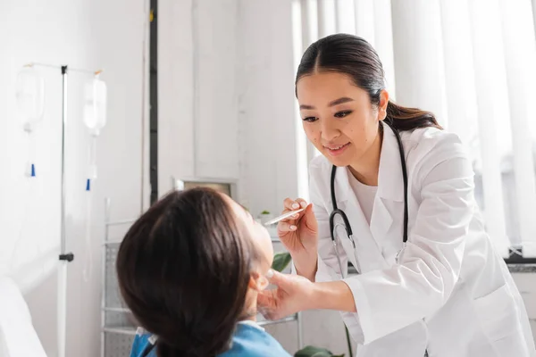 Lächelnder asiatischer Arzt mit Zungendepressor, während er kleine Patientin auf Krankenhausstation untersucht — Stockfoto