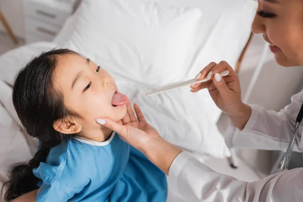 Asiático chica apertura boca cerca sonriente médico con lengua depresor en hospital sala - foto de stock
