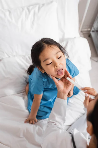 Високий кут зору азіатської дівчини, що відкриває рот біля лікаря з депресором язика в педіатричній клініці — стокове фото