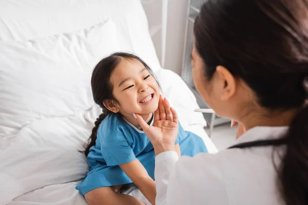 Маленька азіатська дівчинка показує зуби розмитому лікареві з депресією язика в лікарняному палаті — стокове фото